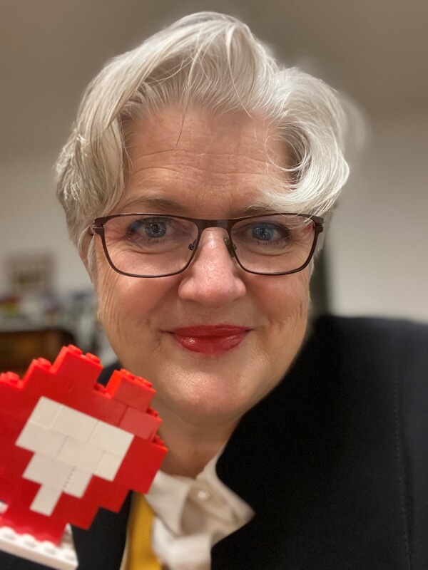 Kopf einer Frau mit Kurzhaarfrisur und Brille , die ein mit roten und weißen Legosteinen gebautes Herz in die Kamera hält