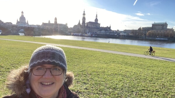 In Winterkleidung gekleidete Frau (Kopf sichtbar)  steht auf einer Wiese, im Hintergrund die Elbe und die Skyline von Dresden