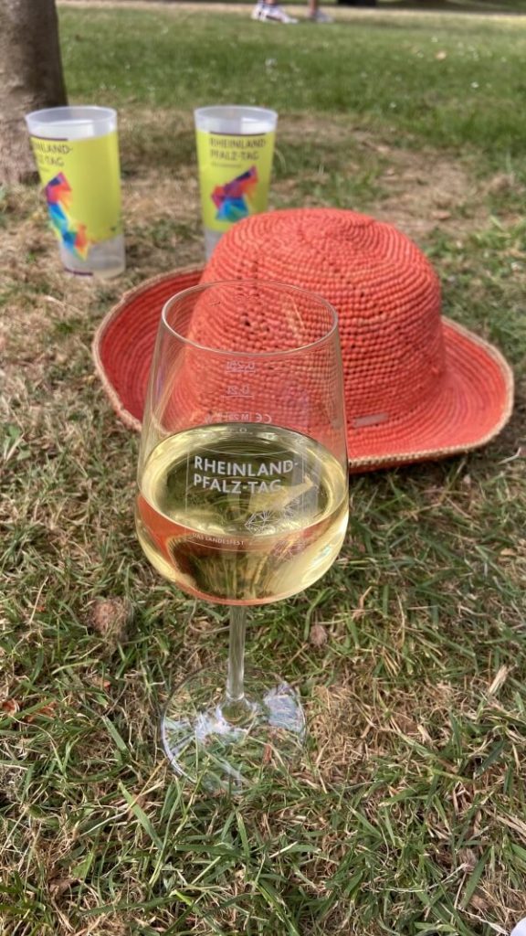 Auf einer Sommerwiese liegt ein apricotfarbener Strohhut; zusätzlichen stehen dazu zwei Plastik-Trinkbecher und eine mit Weißwein gefülltes Weinglas. Auf Letzerem ist zu lesen: Rheinland-Pfalz-Tag