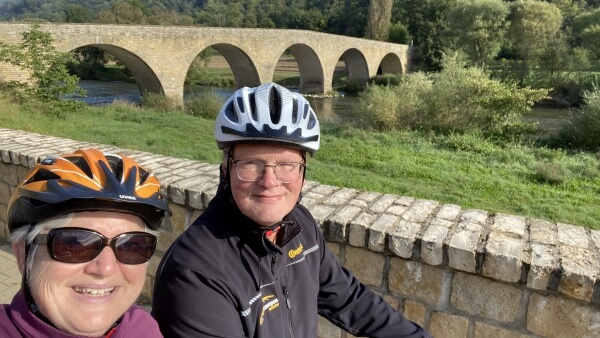 Mann und Frau mit Fahrradhelmen, im Hintergrund eine alte Steinbrücke und ein Fluss